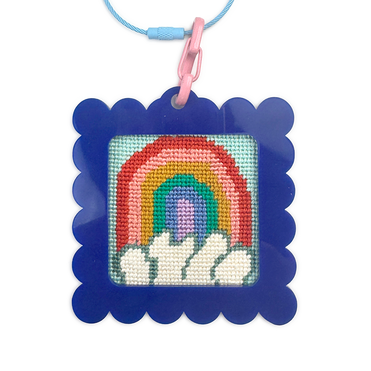 Needlepoint rainbow design in a blue frame with a pink clip on a white background bag tag