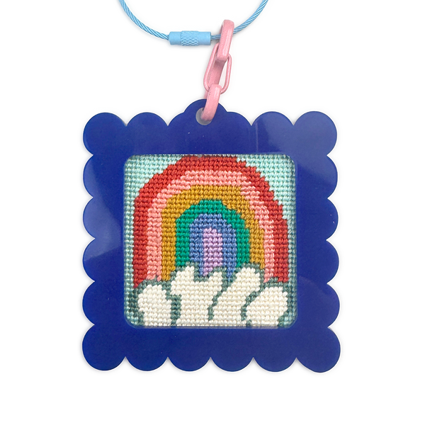 Needlepoint rainbow design in a blue frame with a pink clip on a white background bag tag